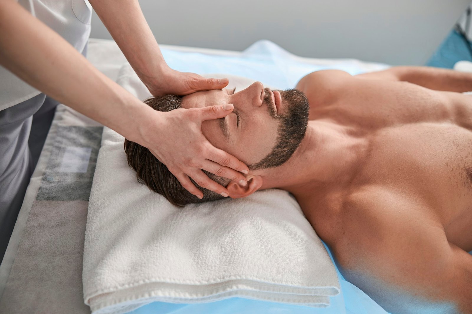 Massage therapist works with head of male lient lying on couch in contemporary clinic